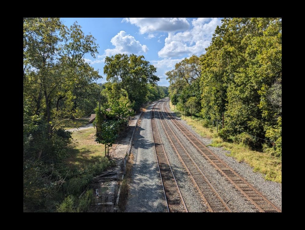 Photo of 3 parallel train tracks receding into the distance with blue skies above. The tracks are lined with trees and there is an abandoned train station on the left.
