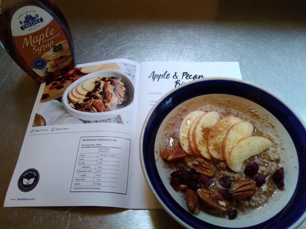 Apple and pecan bircher porridge topped with cinnamon, cranberries and apple slices in a blue and white bowl. A recipe booklet, showing a photo of the same porridge, is held open by the bowl and a bottle of maple syrup.