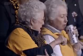 two elderly women are sitting in a stadium watching a game and one is holding a can of beer .