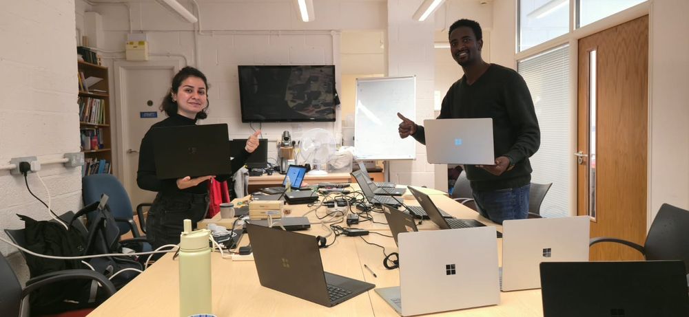 Two smiling Asylum Welcome team members stand beside a long table covered in laptops, each holding one up to the camera to celebrate a donation from Adobe and Revivn.