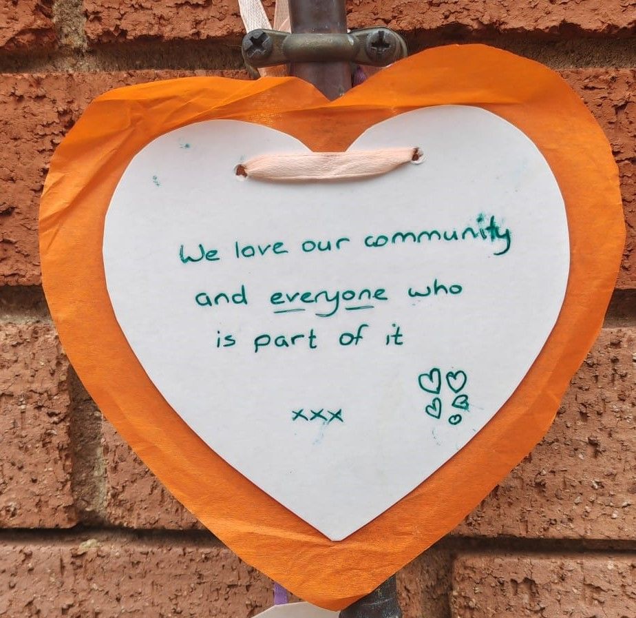 Heart-shaped sign with an orange border and white centre, hanging on a brick wall. The handwritten green text reads, "We love our community and everyone who is part of it xxx" with small heart doodles.