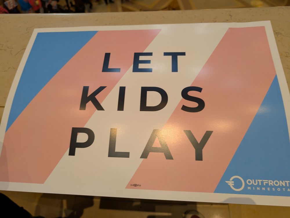 A placard w trans flag background that says "let kids play"
