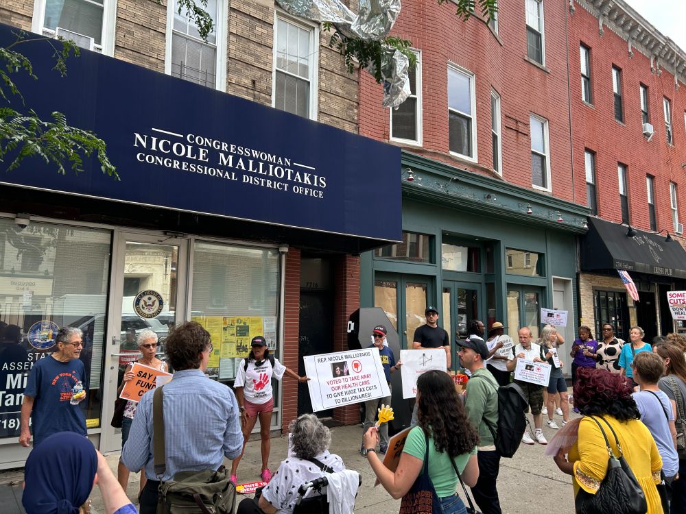 Group of people holding signs gathered on a sidewalk outside of Rep. Malliotakis office.