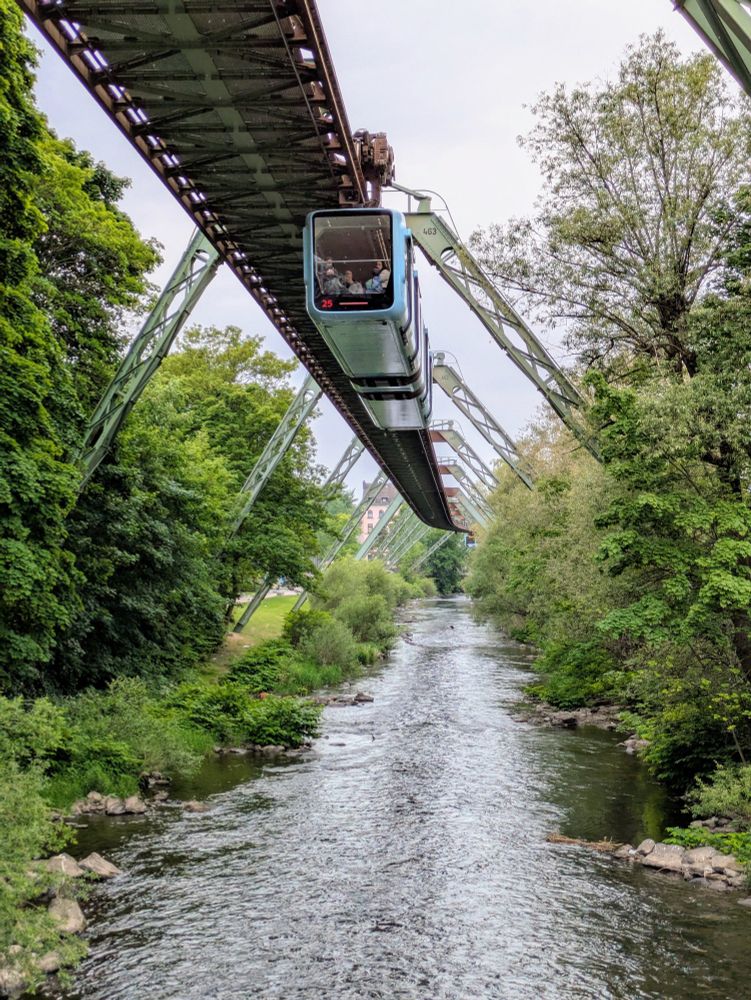 Vue sur la rivière Wupper depuis un pont. Au-dessus s'élève le train suspendu de l'agglomération de Wuppertal. Le rail repose sur une série d'arches qui enjambe la rivière. Sur cet rail circule un train. Composé de deux modules connectés par un "accordéon". Le train est bleu ciel pastel. Il a une allure très moderne qui tranche avec le côté rétro de la structure métallique sur laquelle il circule. Les rives de la rivière sont très fournies en végétation luxuriante. 