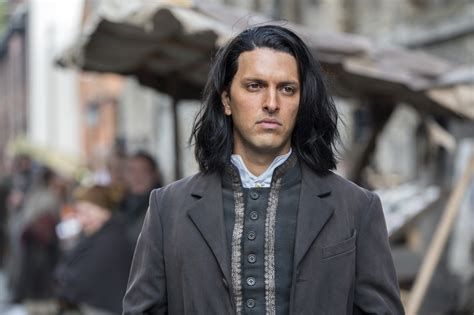 Shazad Latif in the show PENNY DREADFUL. He's a handsome brown man with long dark hair, parted to the side and hitting just at his shoulders. He's wearing a period costume, a gray coat over a high necked vest with gold trimming along the edges and buttoned up to his throat. Underneath that he has a white, high collared shit on. 
