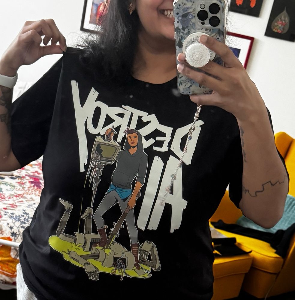 A picture of me with most of my face out of frame (tho you can see I have a big grin on) wearing a shirt that says “DESTROY AI” with a illustration of a person holding a baseball bat with nails coming out of it in one hand, and in the other a broken monitor with bolts dangling down styled to look like a beheaded skull and dangling vertebrae