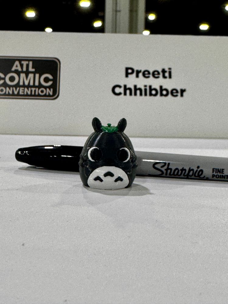 A tiny round Totoro figurine pictured next to a sharpie for size. The sharpie on its side is about half the height of the figurine, which probably also has the diameter of a quarter. You can also see my ATL Comic Con paper name placard behind it.  