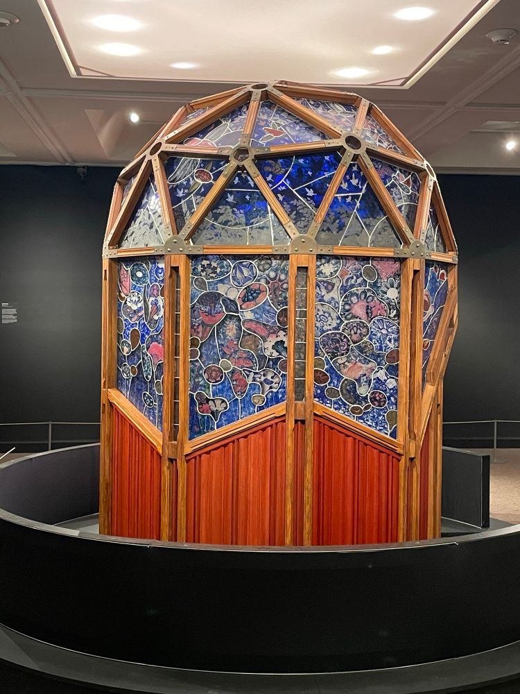 The outside of the pod. It’s 8 feet high and 6 feet wide and finished with wooded panels on the bottom, topped with panels full of fantastical plant and animal life and a geodesic dome of blue. 