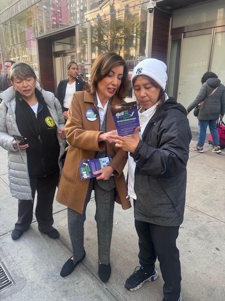 On the sidewalk, Governor Hochul explains the Yes on Affordable Housing ballot props to someone wearing a Yankees hat
