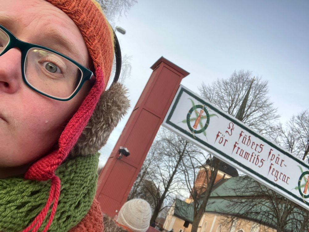 saylo (iförd grön halsduk och röd/brandgul/gul mössa, samt brunlurviga öronmuffar) vid målgången för Vasaloppet, framför mållinjen där det står ”I fäders spår - för framtids segrar.” Den frakturstil som används gör lite så att det ser ut att stå ”J fäder5 fpåc - för framtid5 fegrac.”