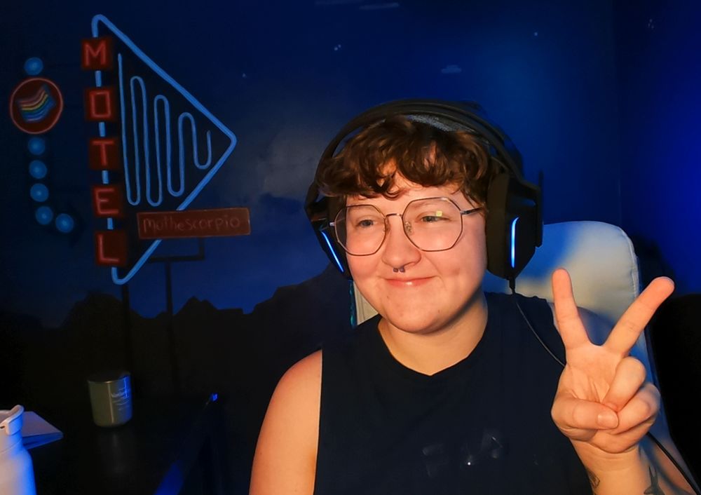 a photo of mo smiling softly looking to the side of the camera while holding up a peace sign. they have a navy tank top, black headphones, black round glasses, and a silver septum ring on. the background is a custom painted mural by their partner jordan that shows a neon blue adn red motel sign that also says "Mo The Scorpio" with a rainbow flag also incorporated into the sky. mo's lavender water bottle is to the left as well as an unlit grey/green candle