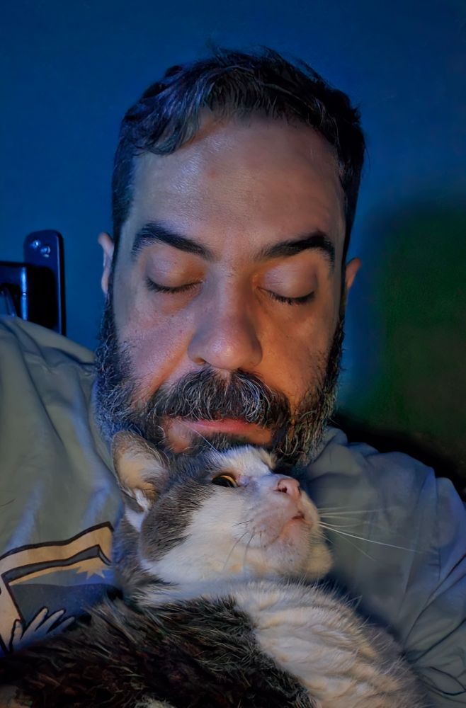 Man sitting up in bed with a cat leaning up against him. 