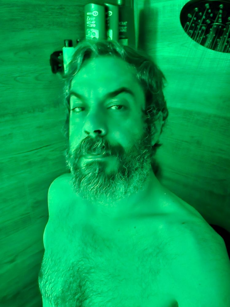 Naked man in the shower. Lit all in green he is visible from mid chest up. He attempts to smile but it is fucking early. 