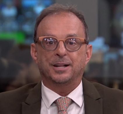 homem de meia-idade usando óculos com armação degradê entre bege e cinza, terno cinza escuro e camisa branca com gravata estampada