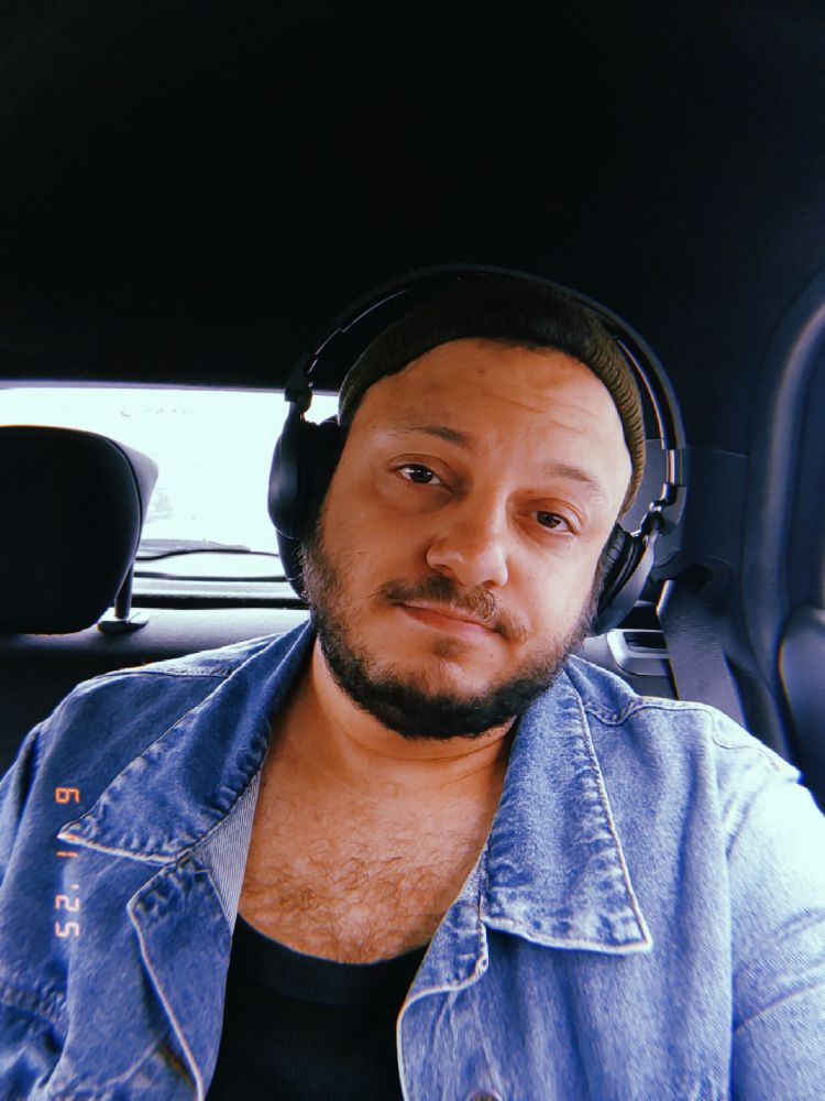 um homem no banco de trás de um carro usando fone de ouvido, gorro verde, jaqueta jeans e camiseta preta 