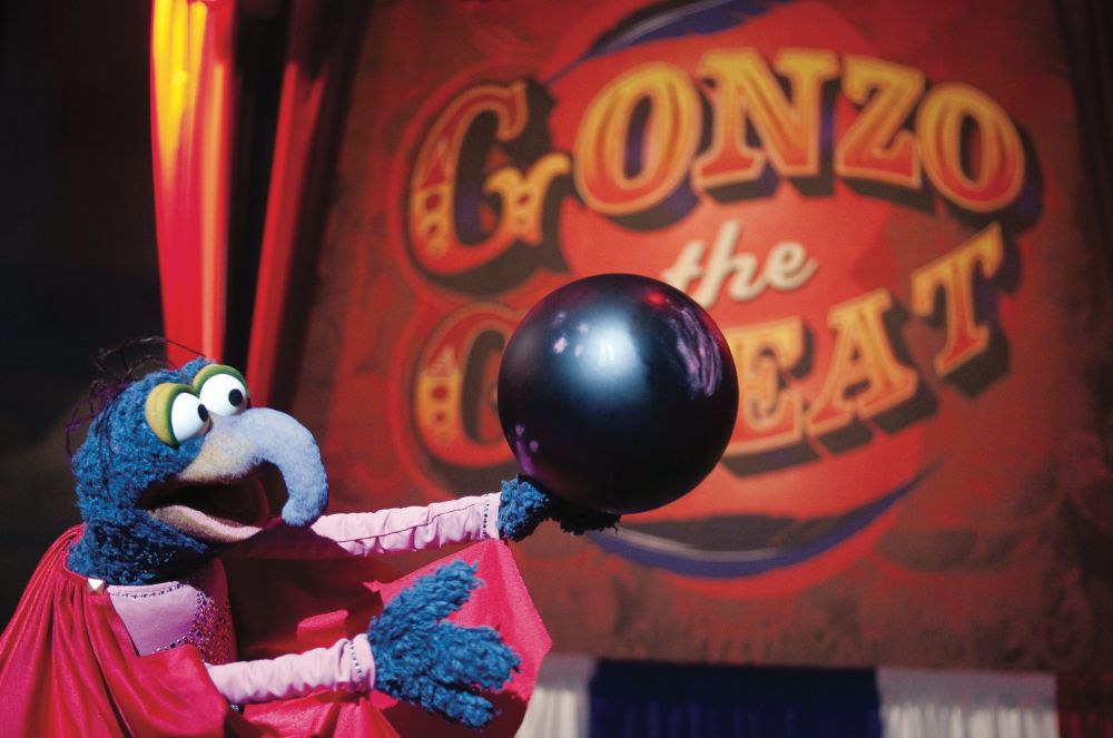 ID: Gonzo the Great wears a pink cape and holds up a heavy looking black bowling ball as if by magic with a red and orange sign in the background that reads "Gonzo the Great".