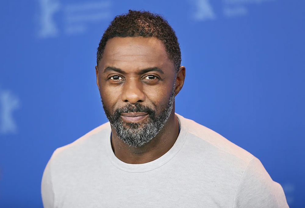 ID: Idris Elba looks directly at the camera with a powerful stare while wearing a white shirt and against a blue background