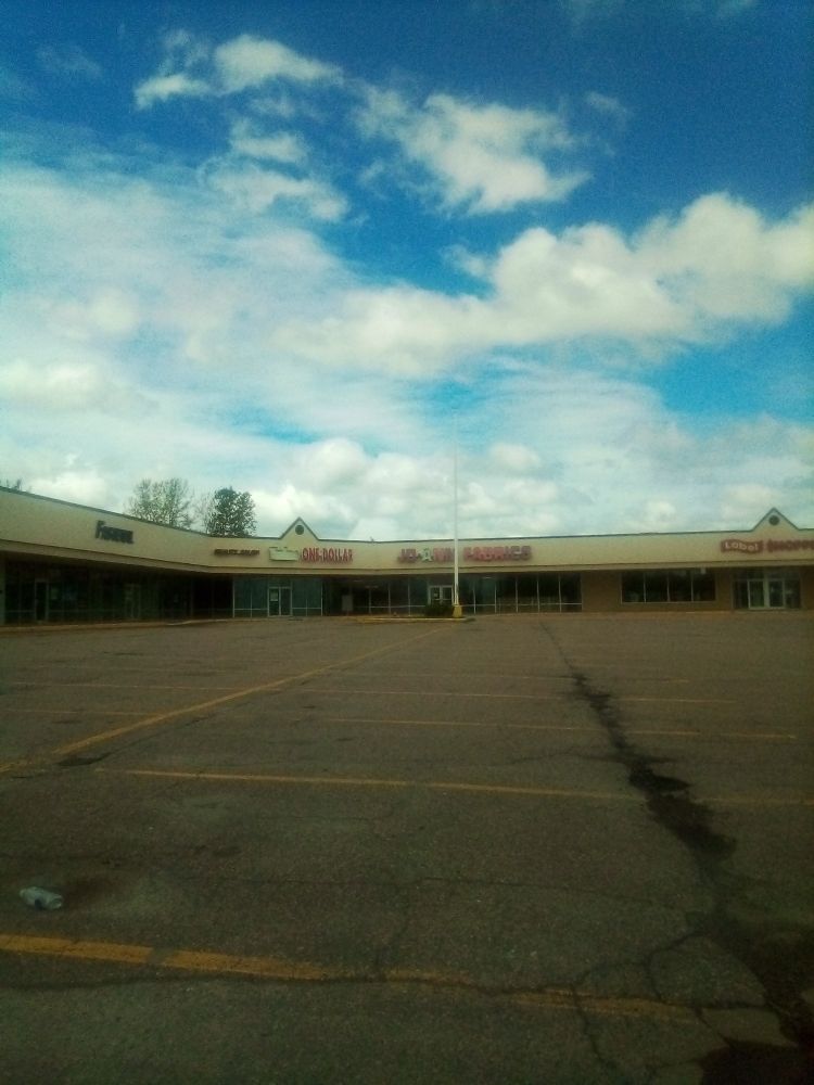 A beautiful fluffy cloud blue sky over a run down shopping center and parking lot. There's a closed Fastenal, Salon, Dollar store, and Jo-Ann fabrics.