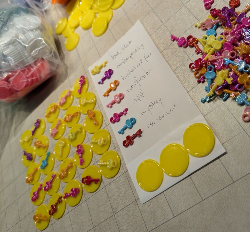 A game board under development. There are yellow discs arranged in a 5x5 grid and topped with miniature skeleton keys in assorted colors. A chart matches the key colors to genres. Additional game pieces are piled nearby.
