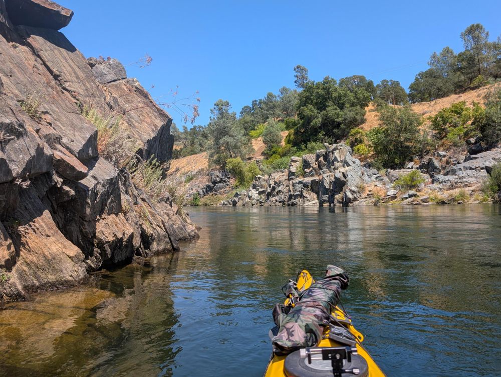 The nose of my yellow kayak with the rocky shores of the river ahead. My bike battery is in a camo dry bag on the kayak's bow.