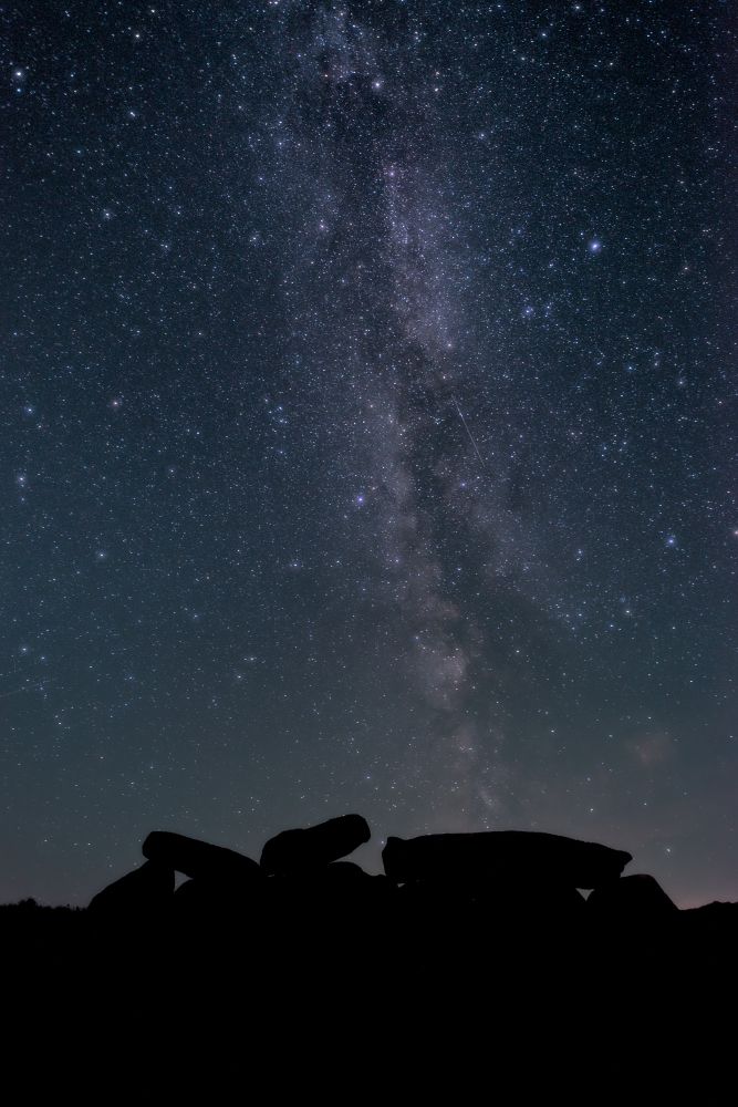 Photo astro de la voie lactée au dessus d'un dolmen qui se détache en avant-plan en ombre chinoise dans les lumières de la côte.