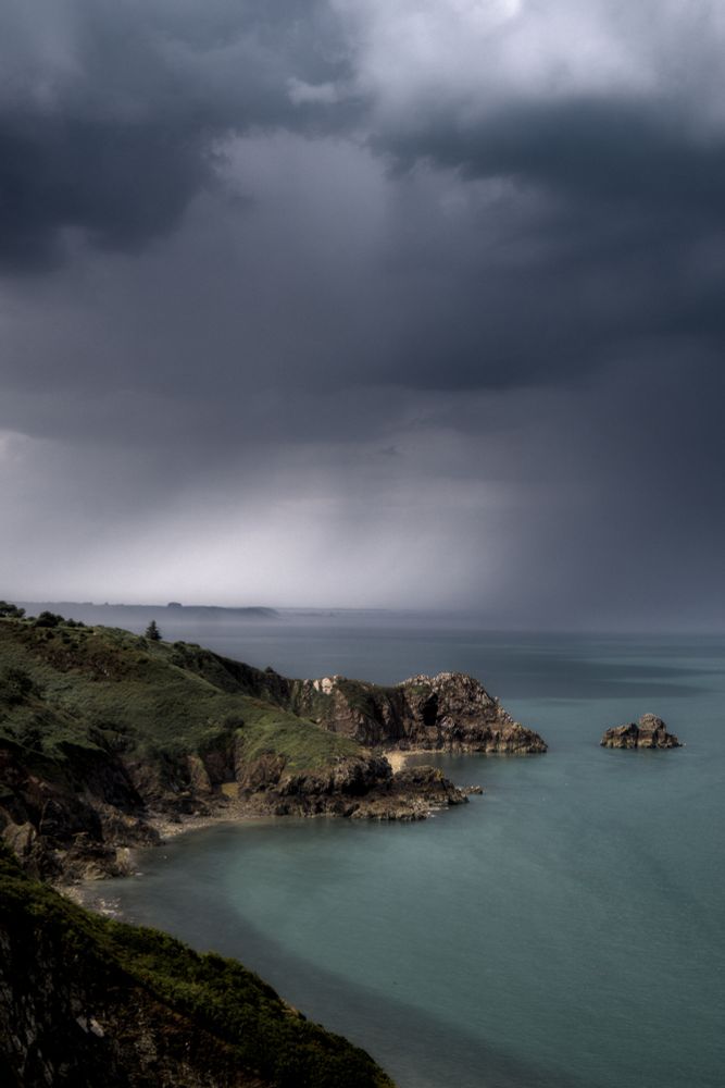 Photo de la côte Bretonne depuis les hauteurs des falaises de Plouha, au loin la mer est prises dans les trombes de pluie d'un orage aux tons bleux glacés et violets alors que près des falaises la mer est turquoise