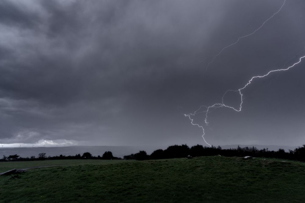 Photo de type paysage donnant sur un gros nuage très sombre et un bel éclair sur la droite de l'image avec de nombreuses ramifications. Sur la gauche on voit des éoliennes sur une crête de collines au loin dans la lumière.