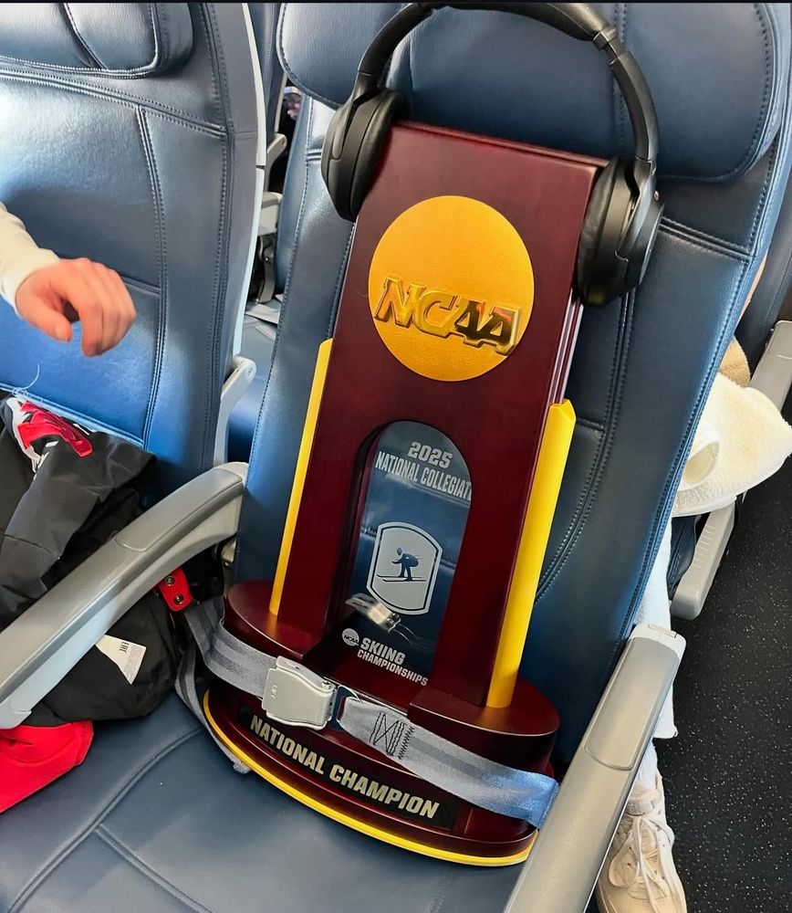 Photo of the NCAA Ski Championship Trophy buckled into an airplane seat with headphones on 