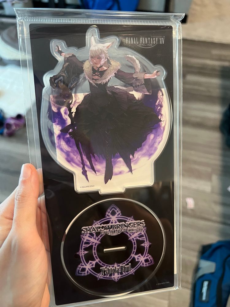 A photo of me holding my new Yshtola shadowbringers acrylic standup!!!!!! It’s mostly black with purple accents, and an overall circular shape 