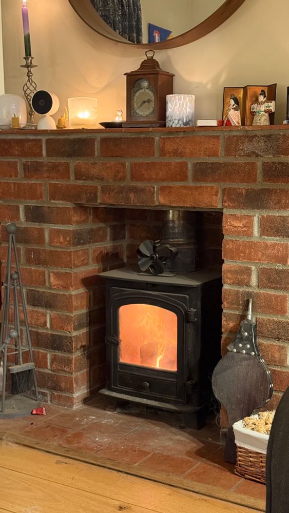 A fire burning in a black iron log burner, the surroundings are terracotta tiles and brick. On the mantelpiece are candles, a clock and Japanese clay figurines. 