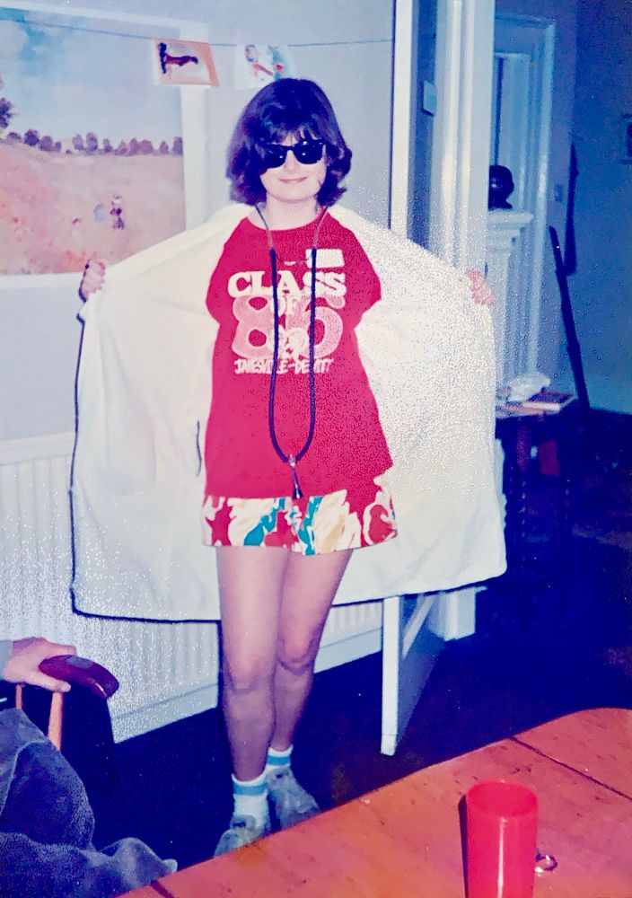 A scan of a photo of a 17 yr old me wearing bright shorts, a class of 86 t-shirt, dark glasses, a stethoscope and a white lab coat. 