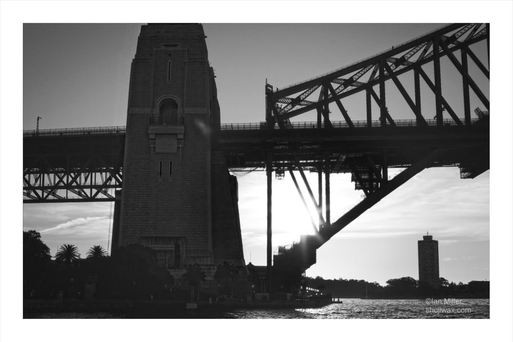 A black and white image of a bridge, featuring a prominent stone tower and a steel structure. The sun is positioned behind the bridge, creating a silhouette effect. In the foreground, there are trees, and a tall building is visible in the distance.