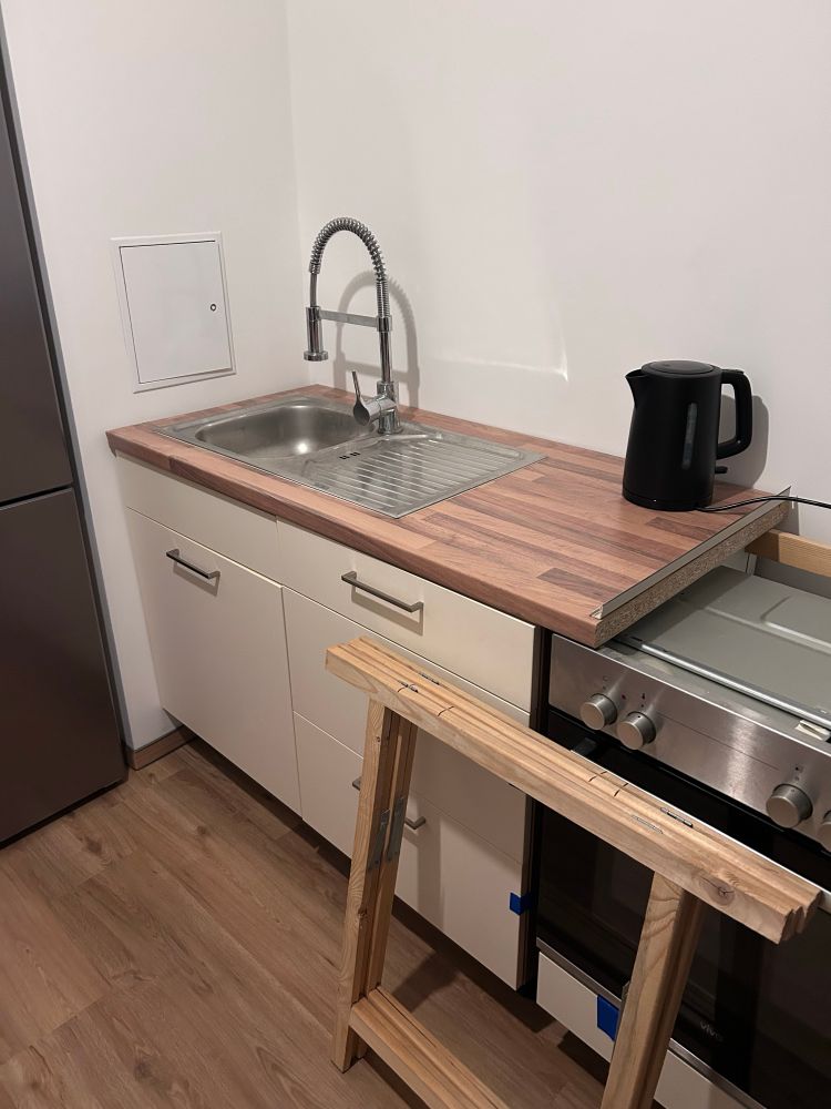 Eine provisorische Küchenzeile die links mit Spülschrank beginnt, dann Schubladenschrank, dann ganz rechts der Backofen. Auf dem Spülschrank und Schubladenschrank liegt eine Arbeitsplatte mit Spülbecken auf. Neben dem Spülbecken steht ein schwarzer Wasserkocher. Am Backofen lehnen zwei Böcke an. 