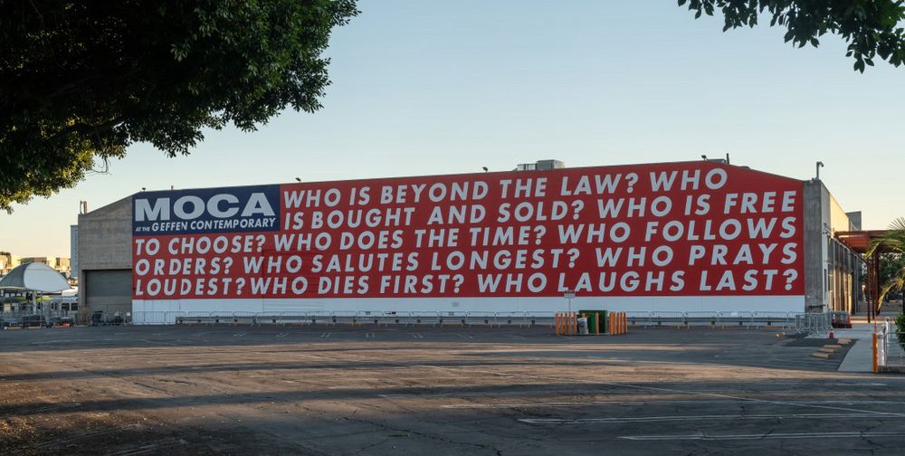 Photo of a Barbara Kruger installation at the MOCA Geffen in LA.  The text (in all caps with white letters against a bright red background) poses the following questions: WHO IS BEYOND THE LAW? WHO wIS BOUGHT AND SOLD? WHO IS FREE
TO CHOOSE? WHO DOES THE TIME? WHO FOLLOWS ORDERS? WHO SALUTES LONGEST? WHO PRAYS LOUDEST? WHO DIES FIRST? WHO LAUGHS LAST?