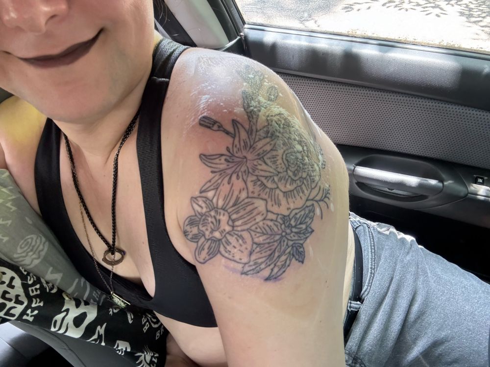 A woman in a sports bra, dark lipstick, and gray jeans showing off a picture of a fresh tattoo covered with Second Skin tattoo cover. The tattoo is linework of botanicals that have a connection with the Greek underworld and death; asphodel, narcissus, mint, cypress, and a rose