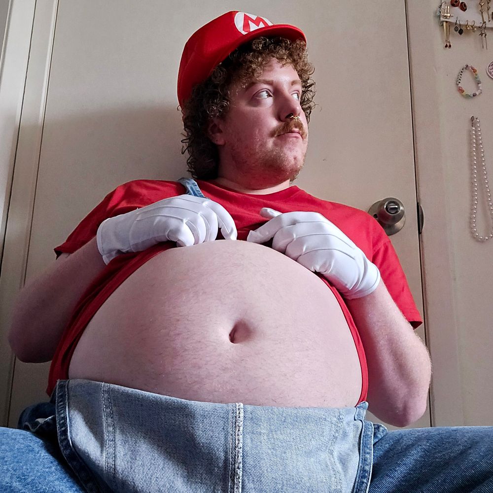 I'm wearing a Mario costume, and sitting facing the camera while I'm looking to the side. My hands are gloved and are pulling back my red shirt while my overalls are unhooked, exposing my round belly.