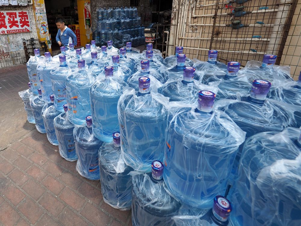 Large blue transparent plastic water bottles (the kind used in water dispensing machines), each covered in a thin layer of clear plastic wrapping, are stacked on a street corner in China. The water bottles dominate the photo frame as the main subject. In the background, light brown and yellow buildings can be seen. A child wearing a blue school uniform walks past, and Chinese writing is visible on a nearby wall.