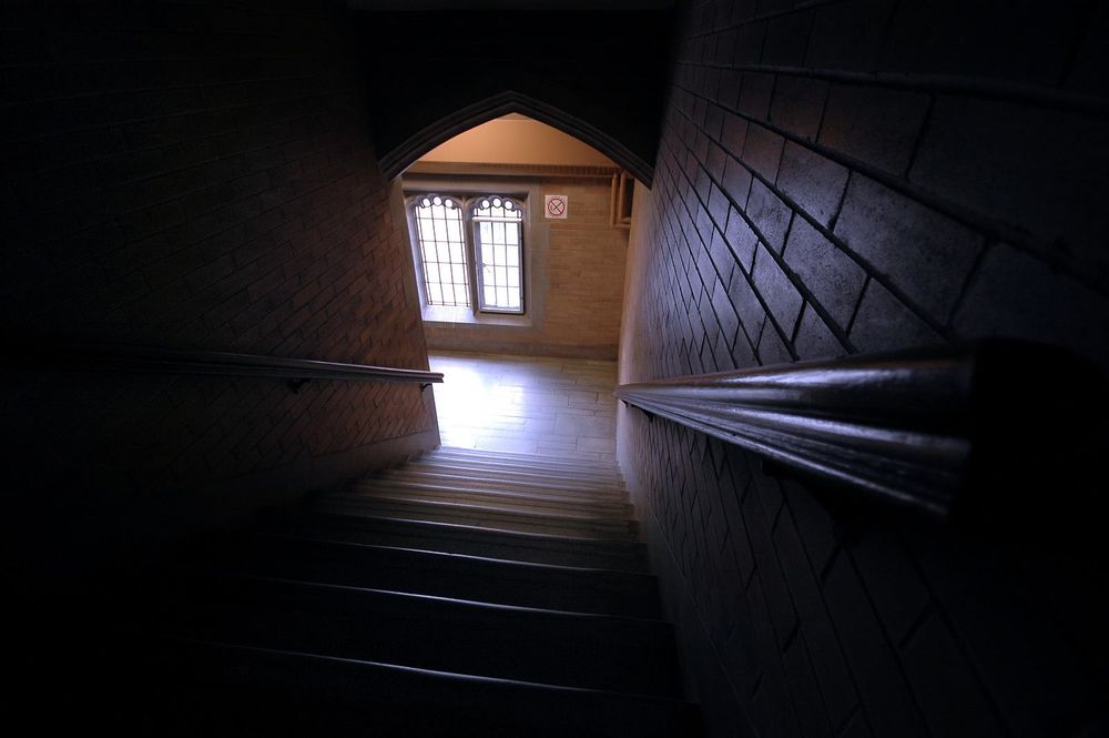 Photograph taken looking down a dark staircase lit at the bottom by a window. 