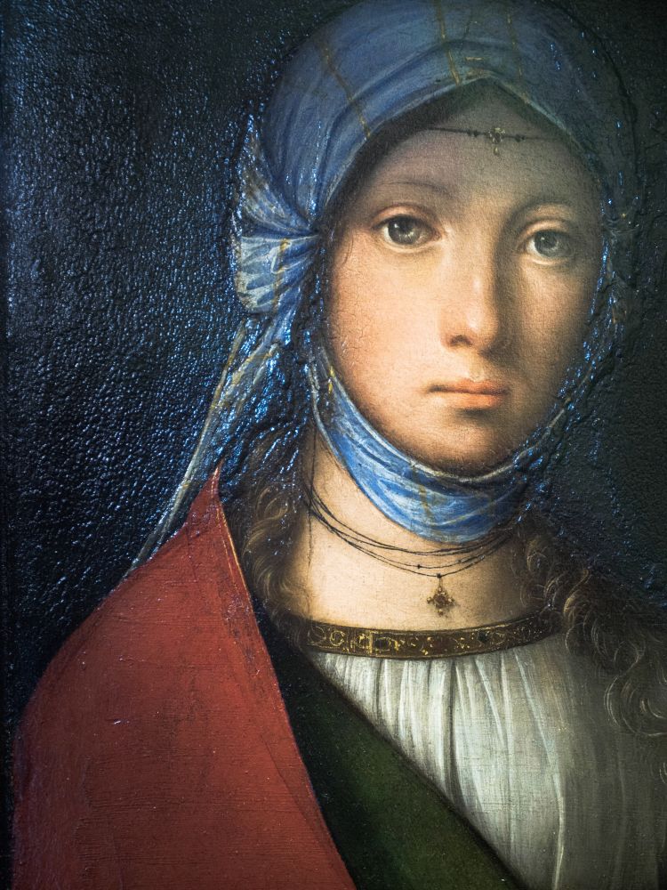 Painting of a young woman known as the "Gypsy Girl" by Boccaccio Boccaccino, circa early 16th century. The half-length portrait shows a dark-haired girl with large, expressive eyes, wearing a blue headscarf, a red cloak, and a white blouse—colors traditionally associated with the Virgin Mary symbolizing purity (white), divine love (red), and heavenly grace (blue). Her gaze is turned slightly to the side, with a serene yet introspective expression typical of Marian portraits. The dark background emphasizes the softness of her features and the luminous quality of her garments.