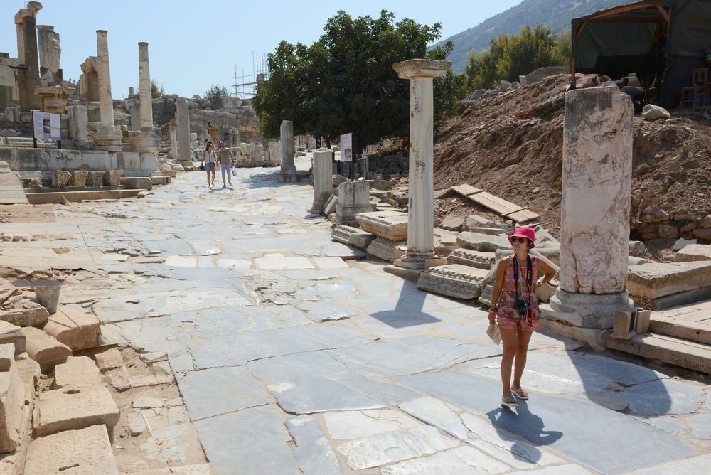 An ancient street paved with work stone with the ruins of buildings on each side. In the foreground is  a woman looking around her, in the background further up the street there is a couple.