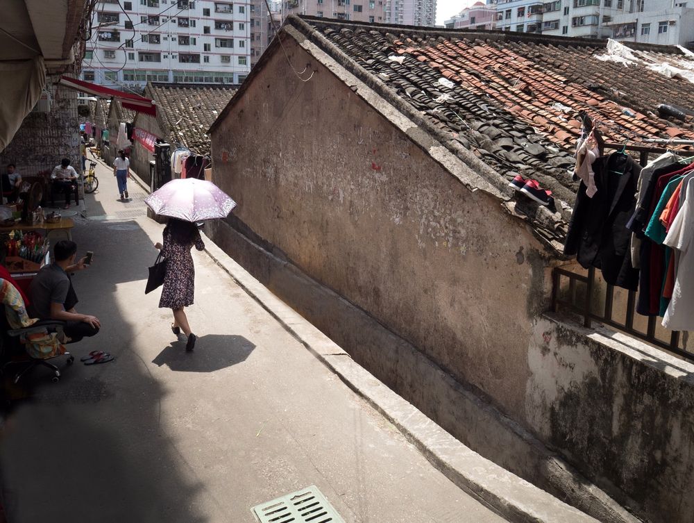 Photo looking down a narrow sunny street with an old tiled-roof building on the right. A woman walks in the middle left holding an umbrella to shield herself from the midday sun—the woman is Chinese, but her face is not seen, so her identity is a mystery. Though not literally Mary Poppins, the scene suggests that idea. Using “Mǎlì Bōpíngsī” in the caption privileges a Chinese take on this Western theme and adds to the mystery for those who do not read Simplified Chinese. 

照片俯视一条狭窄阳光明媚的街道，右侧有一座带瓦顶的老建筑。一名女性走在街道中偏左的位置，撑着伞遮挡正午的阳光——她是中国人，但面容未露，身份成谜。虽然她并非真正的“玛丽·波平斯”，但画面让人联想到这个形象。标题中使用“玛丽·波平斯”体现了对这一西方主题的中国化解读，也增加了不识简体中文读者的神秘感。