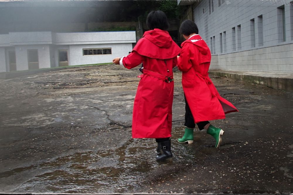 Two women walk side by side in the rain, wearing bright red raincoats. 