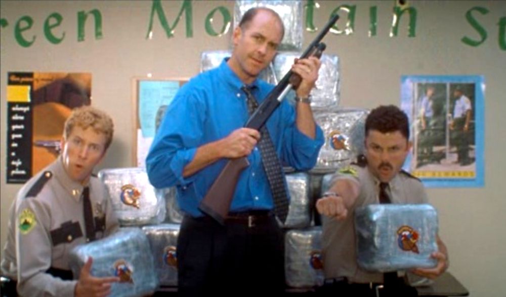 Paul Soter, John Bedford Lloyd and Steve Lemme in a scene from Super Troopers (2001) where they are posing in front of a tower of blocks of The Reefer with a Johnny Chimpo monkey logo on each block. Lloyd who plays the mayor is holding a shotgun hoping to score points with the electorate.