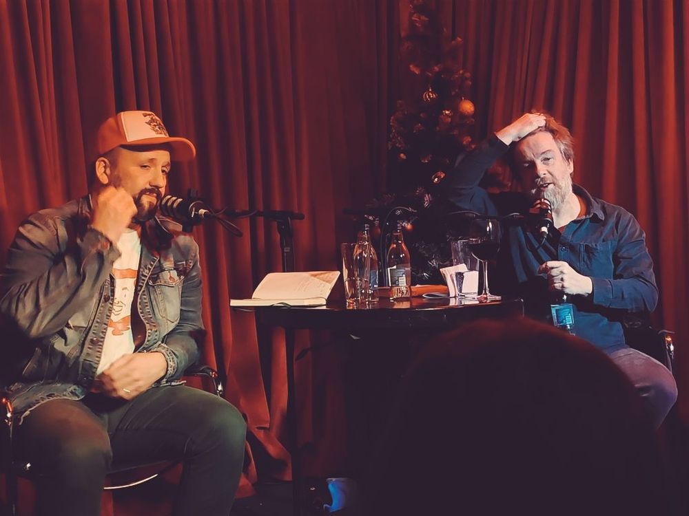 Steve Ryan and Kevin Barry sitting on the stage of the record room with a red velvet curtain behind them
