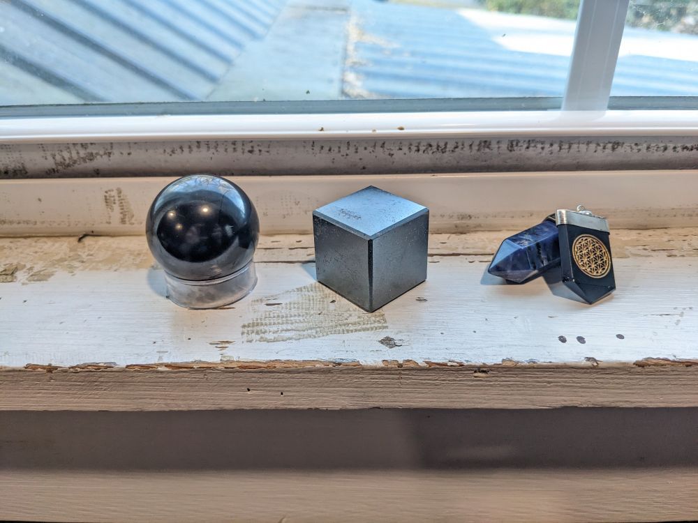 A collection of minerals in front of a windowsill including a grey sphere with semi-lustrous finish, a grey metalloid cube, a pointed hexagonal pendant of blue mineral, and a flat black mineral pendant with the Flower of Life symbol inlaid on it in gold