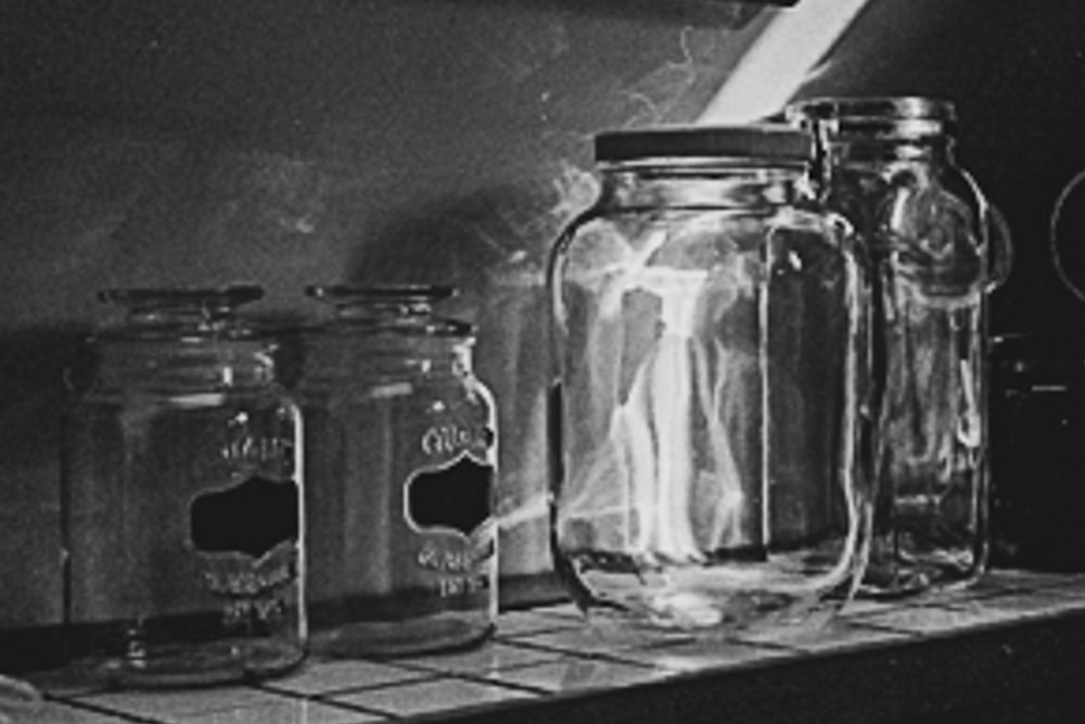 Black and white photograph of 4 empty glass jars on a tile counter and against the wall. 2 smaller and the same, one and old large pickle jar and the 4th a more tall square shape jar. with a sunbeam hitting and refracting in one off the larger pickle jars. Light patterns also reflect on to the wall making interesting patterns. 