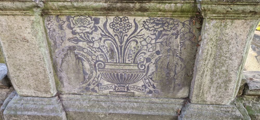 The back of a gravestone with leaded decoration which says 'Music, Arts, Drama'. The name has worn off the front. 
