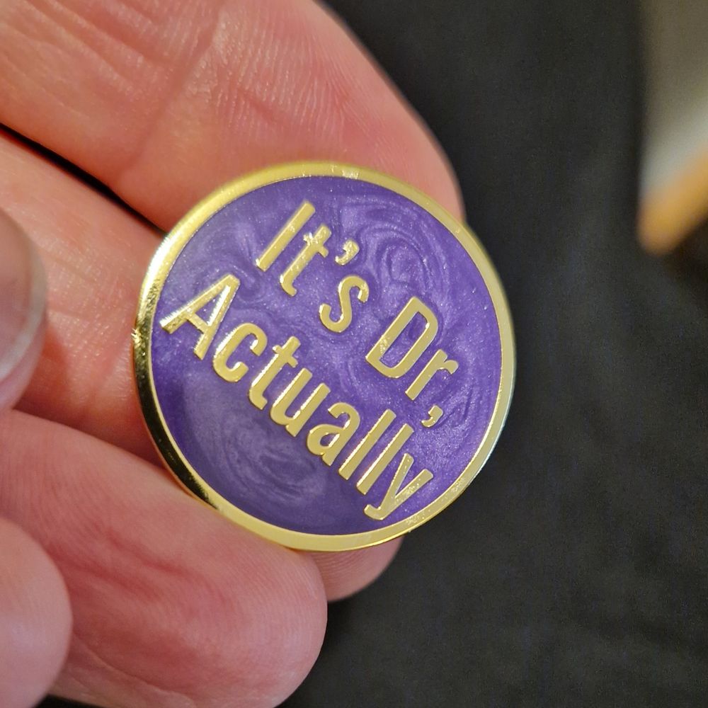 Purple pearlescent soft enamel on that says "It's Dr, Actually".
(Now I want a silly one that says "It's Dr, Acula"...) 