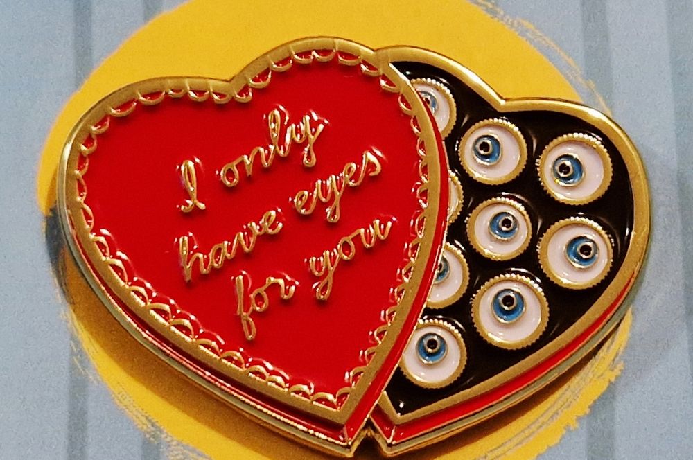 An enamel pin.
Red heart-shaped old-fashioned chocolate box. The open lid has gold decoration and says 'I only have eyes for you' and inside there are cute little eyeballs where the chocolates should be. 