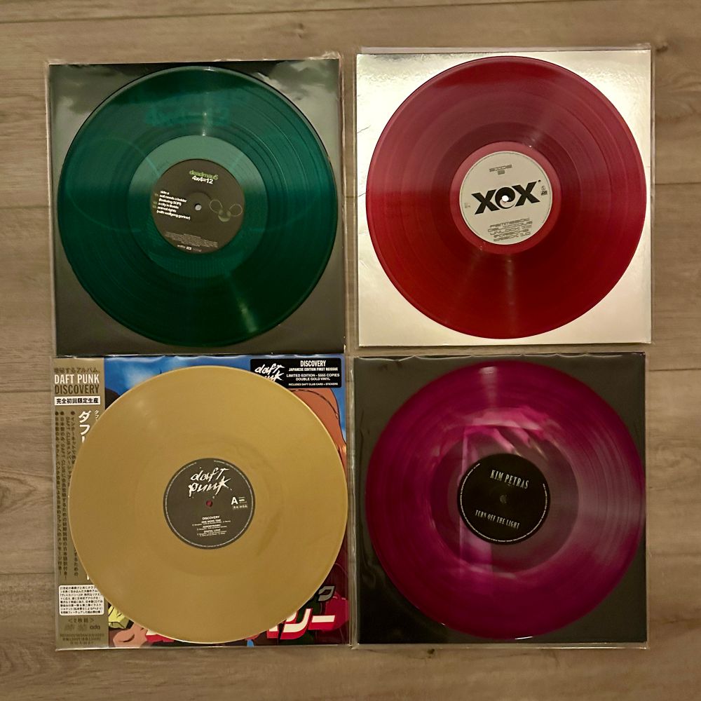 A grid of four vinyl records laid on top of the jackets, a dark green over a black jacket, a dark purple over a white jacket, a gold over a multicolor jacket, and a translucent purple swirl over a black jacket 